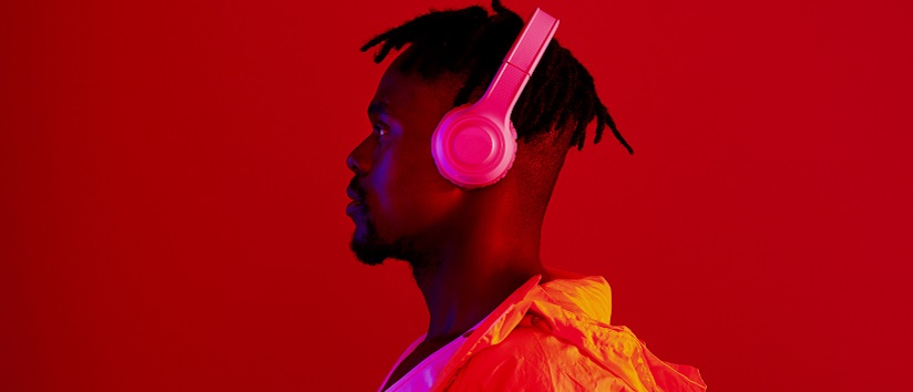 A young black man is standing in profile, he has short dreadlocks and wears pink headphones, he is lit with a red light 