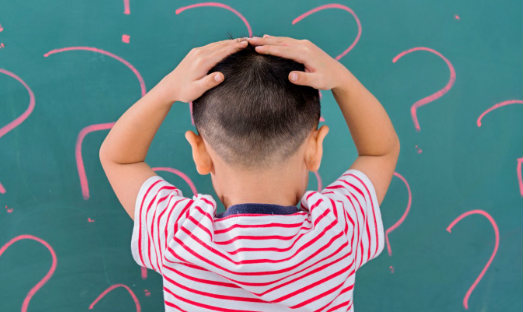 A small child with his back turned and both hands on his head, in front of a blackboard with question marks drawn on it 