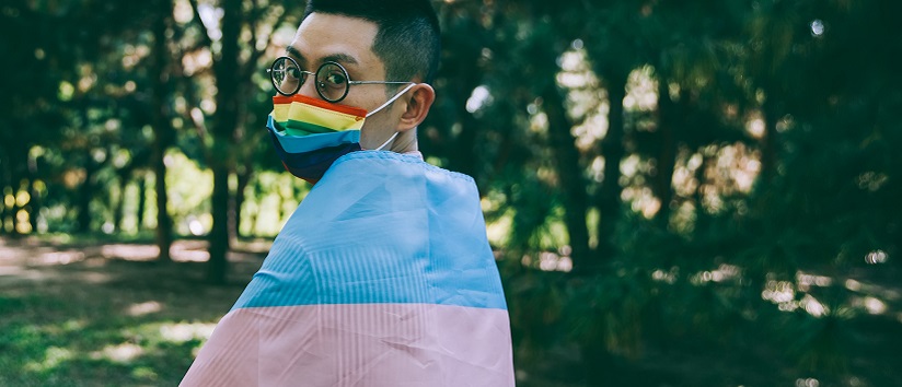 person wrapped in transgender flag wearing a facemask with pride flag printed on it