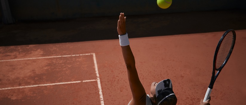 female tennis player is throwing up ball to serve, we see her from behind, she is on an terracotta clay court