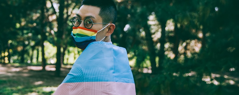 person wrapped in transgender flag wearing a facemask with pride flag printed on it